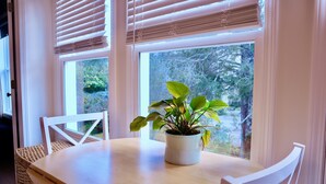 Una mesa de madera con una planta en maceta, sillas blancas y vistas a árboles a través de la ventana.