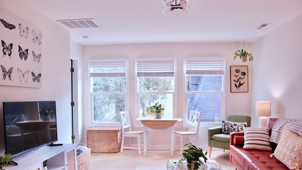 Una sala de estar con televisión, un sofá rojo, un sillón verde, una mesa con sillas y ventanas amplias.