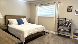 Un dormitorio con cama, mesita de noche, lámpara, ventana con cortinas, estantería y una silla.