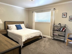 Un dormitorio con cama, mesita de noche, lámpara, ventana con cortinas, estantería y una silla.