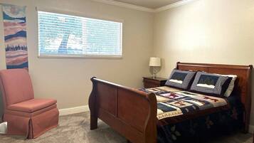 Een slaapkamer met een houten bed, een stoel, een raam met jaloezieën en een muur met een schilderij.