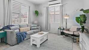 Ein modernes Wohnzimmer mit grauem Sofa, einem weißen Couchtisch und einem beigefarbenen Sofa. Es gibt zwei Fenster mit Jalousien, eine hängende Pflanze und eine Lampe auf einem Beistelltisch.