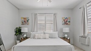 Ein modernes Schlafzimmer mit einem großen Bett, zwei Nachttischen mit Lampen, einer Topfpflanze und Kunstwerken an den Wänden.
