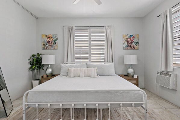 A modern bedroom with a large bed, two bedside tables with lamps, a potted plant, and artwork on the walls.