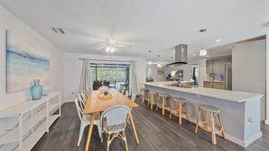 Una cocina moderna con una isla central, armarios blancos y un comedor con mesa de madera y sillas de mimbre.