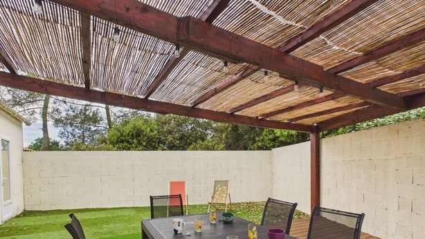An outdoor patio with a table and chairs, a pergola overhead, and a white brick wall in the background. - House with garden, terrace & pétanque - 6 guests (Saint-Hilaire-de-Riez)