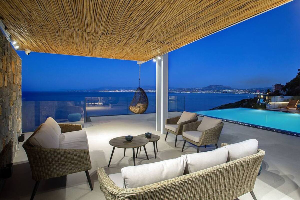 A rooftop terrace with wicker furniture, a hanging chair, and a pool overlooking a cityscape at dusk.