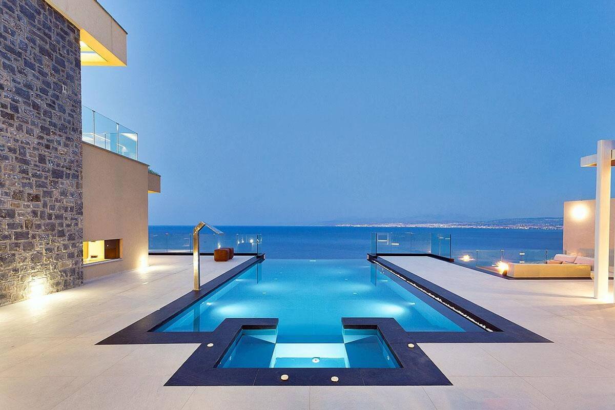 A modern infinity pool with clear blue water, surrounded by a stone wall and a glass railing, overlooking a cityscape at dusk.