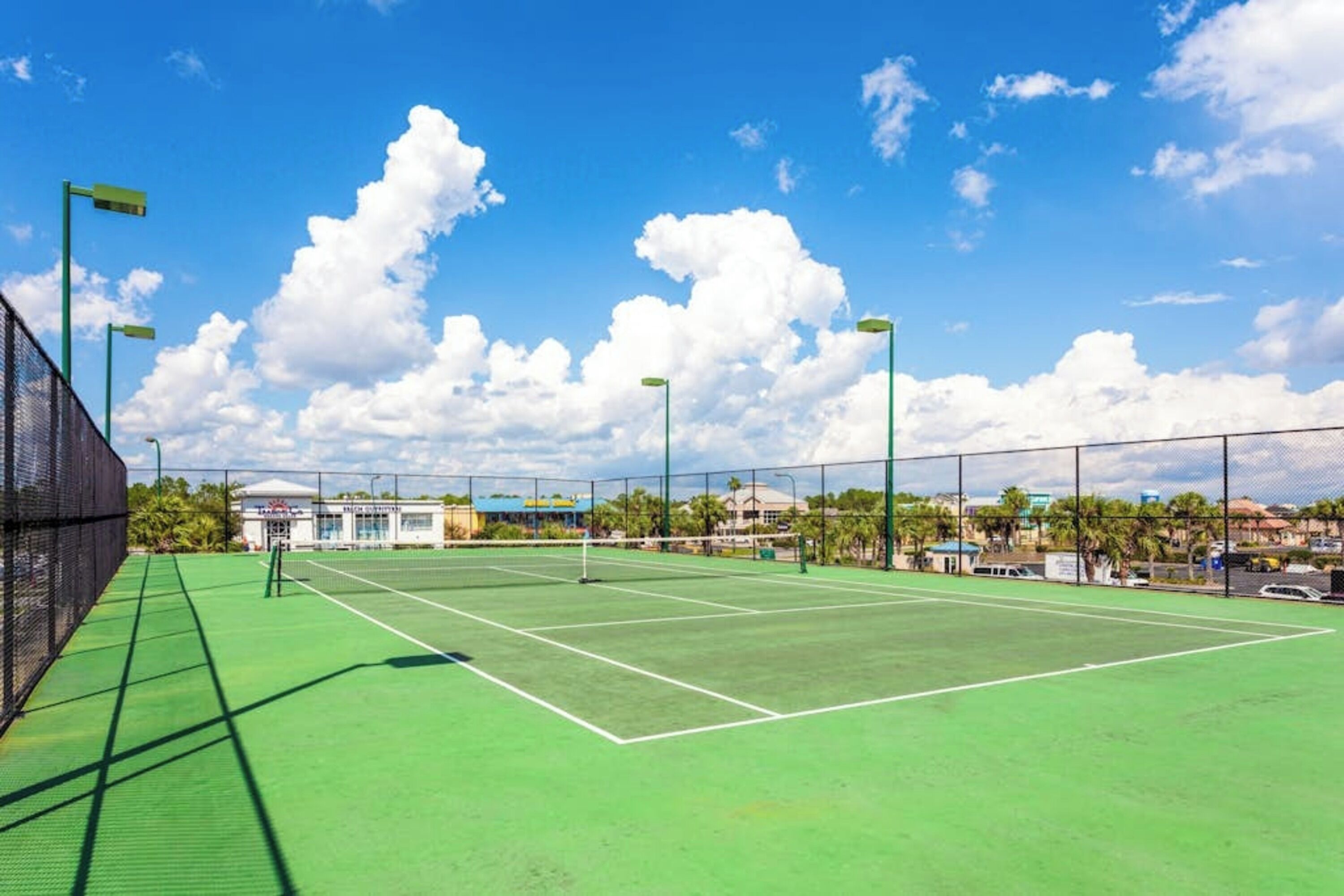 Un court de tennis doté d’une surface verte, entouré d’une clôture, sous un ciel dégagé parsemé de nuages.