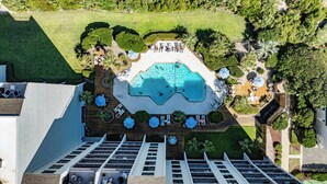 An aerial view of a resort with a central swimming pool, surrounded by lounge areas and greenery.