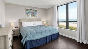 A bedroom with a large window, a bed with a blue bedspread, a nightstand, a lamp, and a painting on the wall.