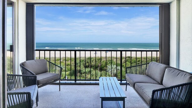 A balcony with two sofas and a table, overlooking a beach with a clear view of the ocean.