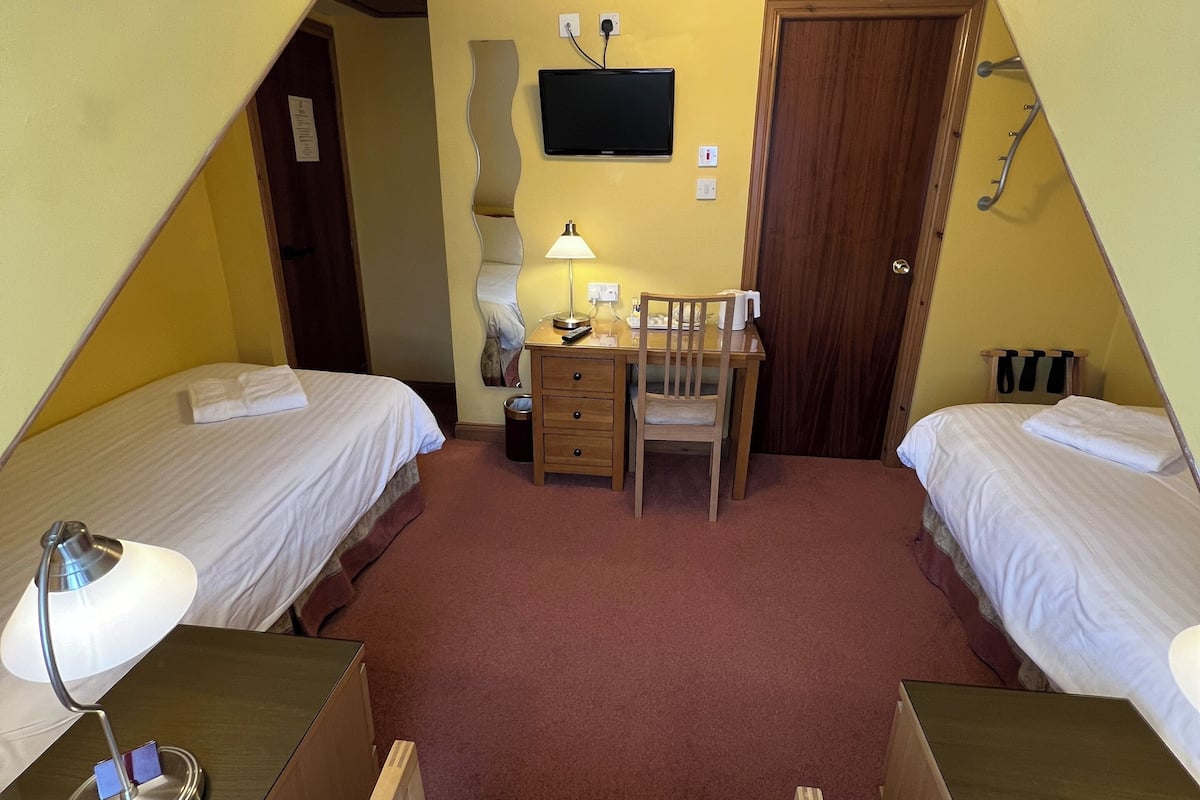 A triangular room with two single beds, a desk with a chair, a television, and a door.