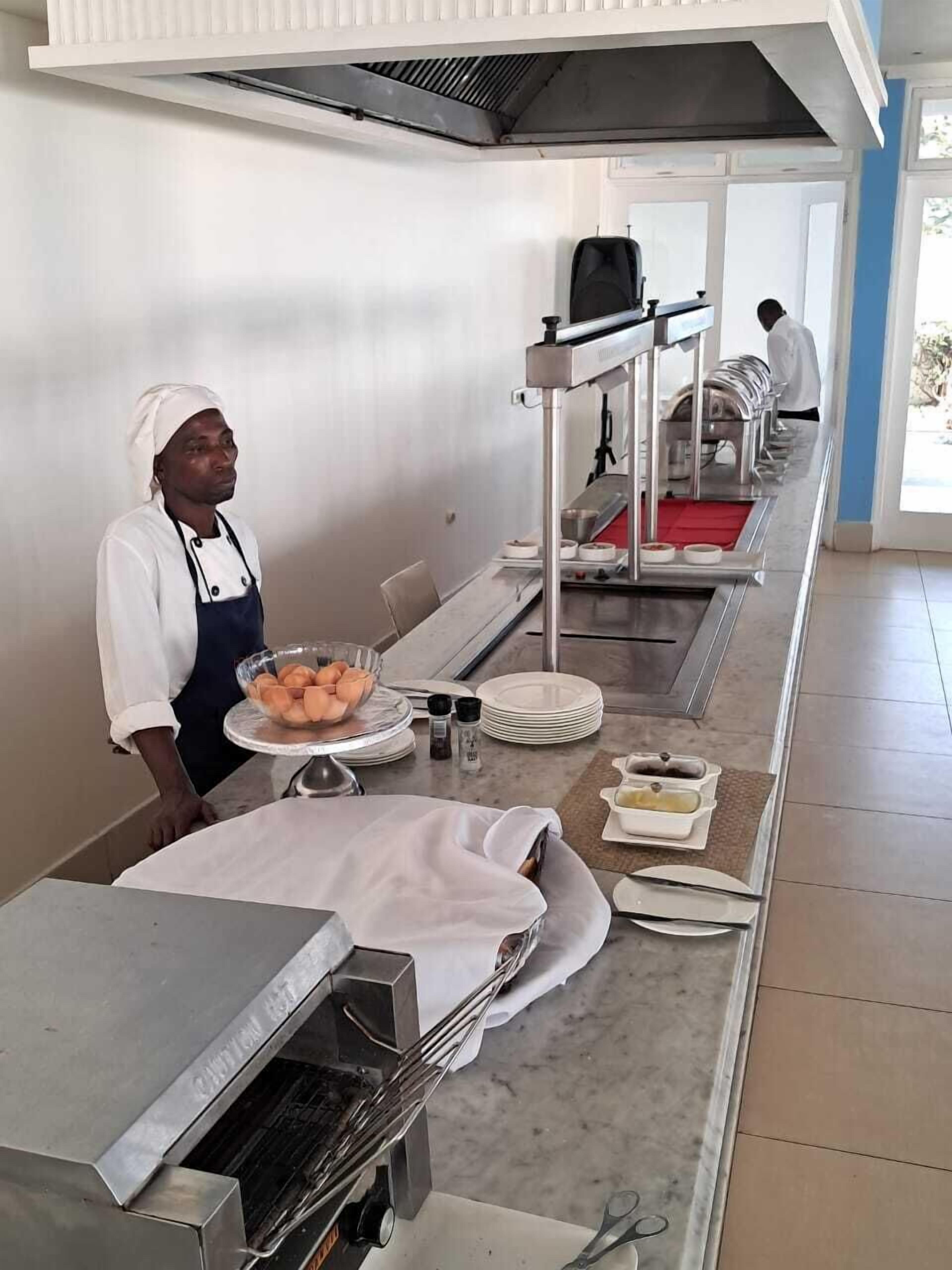 A kitchen area with a chef preparing food, a buffet setup with plates and bowls, and a ventilation hood above.