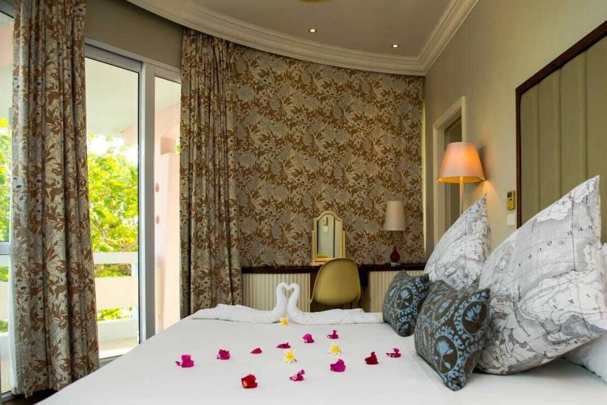 Ein Hotelzimmer mit Bett, floralem Wandpapier, einem Schreibtisch mit Lampe und Blick durch ein Fenster.