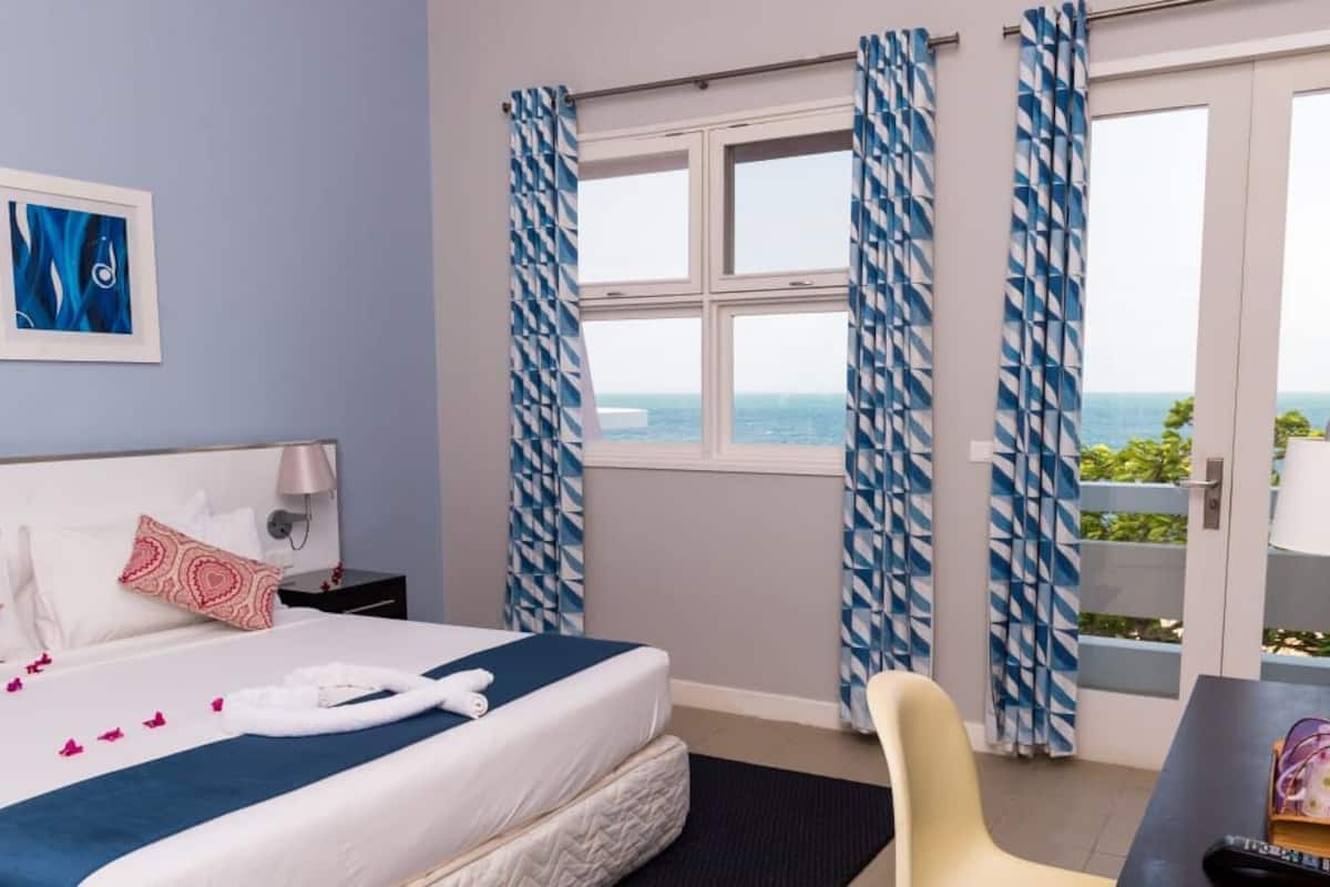 Ein Hotelzimmer mit Bett, Schreibtisch, Stuhl, Nachttisch, Lampe, einem Fenster mit Vorhängen und Blick aufs Meer.