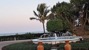A landscaped garden with a central fountain, palm trees, and a view of the sea. - Dona Ana Hotel (Vilanculos)
