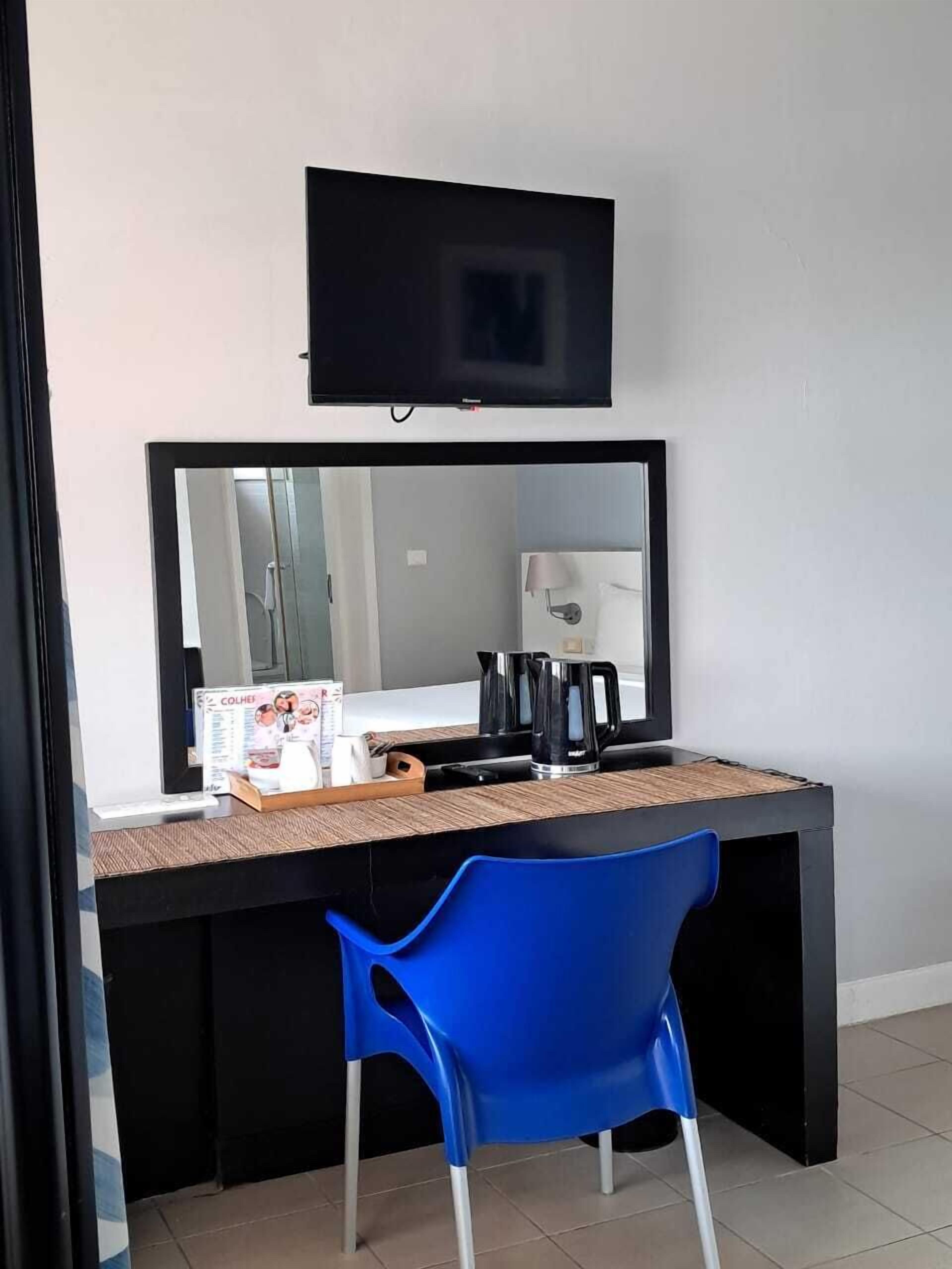 Una habitación con escritorio, silla, televisor montado sobre el escritorio y un espejo que refleja la habitación.