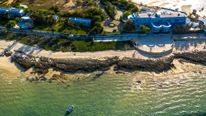 An aerial view of a coastal resort with a beach, a rocky shoreline, and several buildings with blue roofs. - Dona Ana Hotel (Vilanculos)