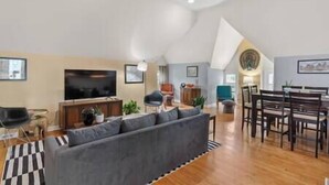 A spacious loft with a high ceiling, a large gray sofa, a dining area with wooden chairs, and a flat-screen TV.