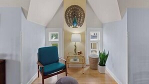 A room with a high ceiling, a blue armchair, a small table with a lamp, and a potted plant.