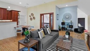 A modern living room with a grey sofa, wooden coffee table, and a striped rug. There is a dining area with a table and chairs, and a kitchen with wooden cabinets.