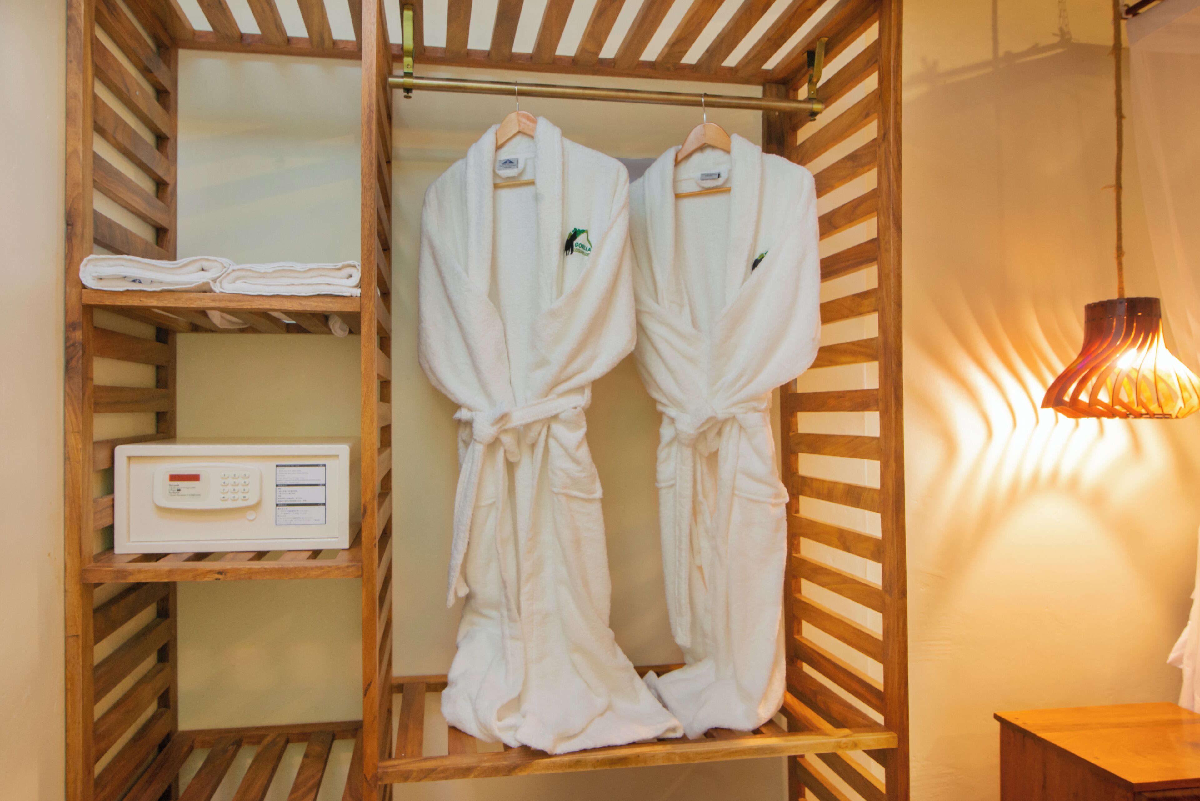 A wooden wardrobe with two white robes hanging on a rack, a white electronic device on a shelf, and a wooden nightstand with a lamp.