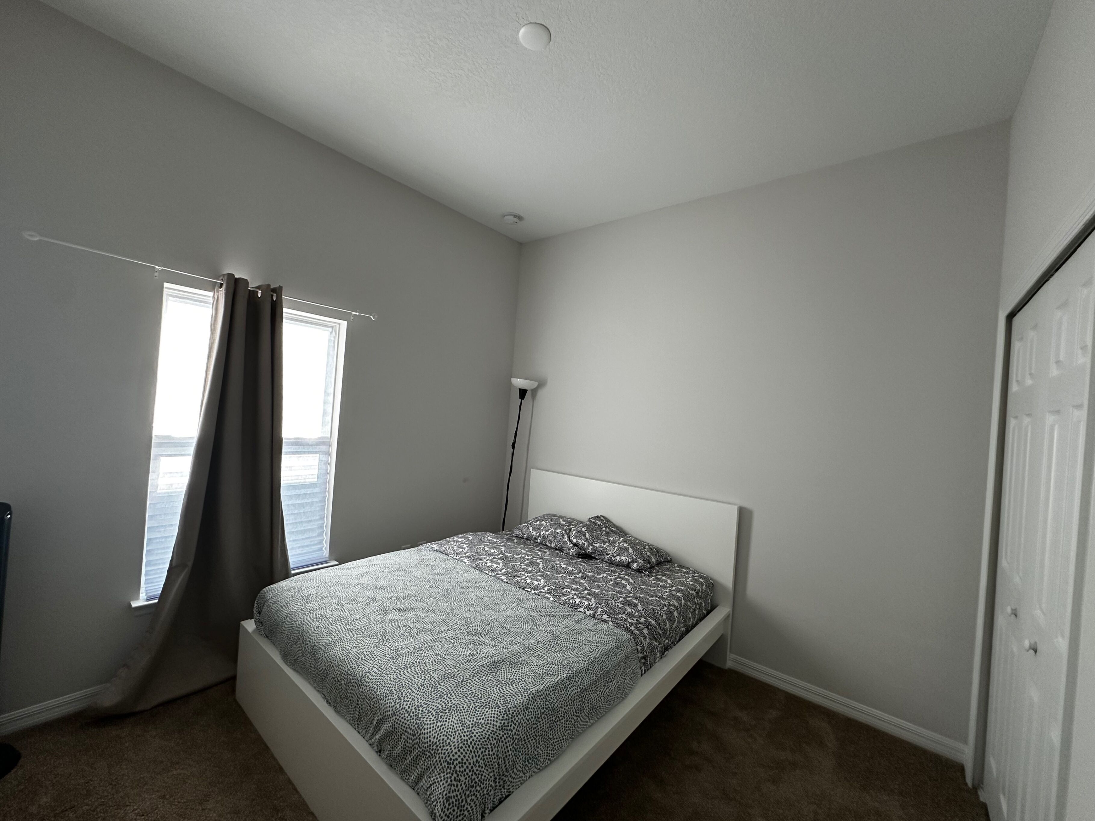 Una habitación pequeña con una cama individual, cabecera blanca sencilla, ventana con cortinas y una puerta.