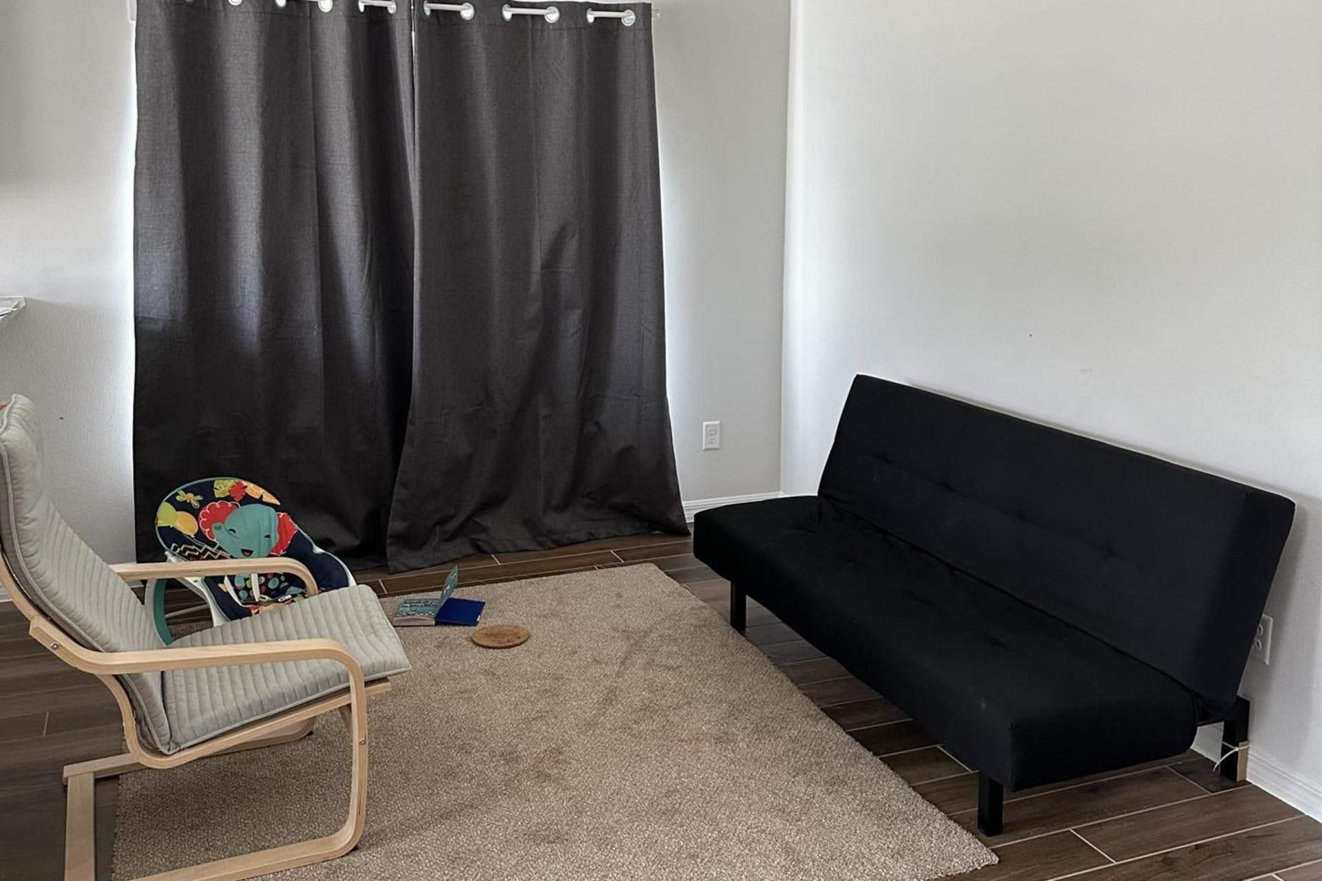 A room with a black sofa, a wooden chair, a rug, and a curtain.