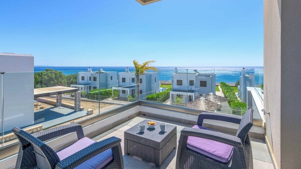 A balcony with wicker furniture and a table, overlooking a coastal view with buildings and the sea.