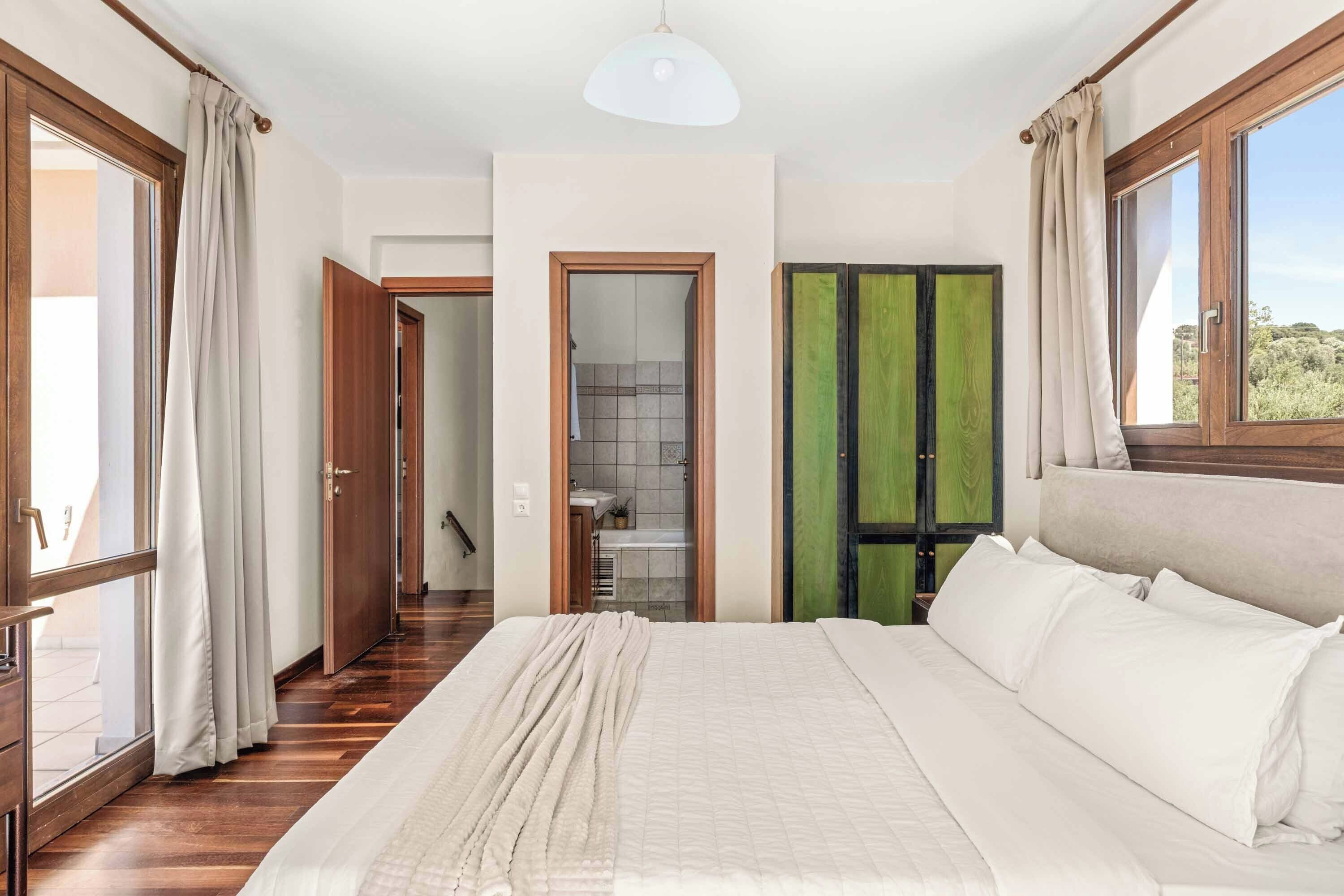 Une chambre à coucher avec un grand lit, un plancher en bois, une vue par la fenêtre et une porte donnant sur la salle de bain.
