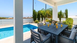 Ein überdachter Essbereich im Freien mit Pool, Sitzgelegenheiten und Blick auf das Meer.