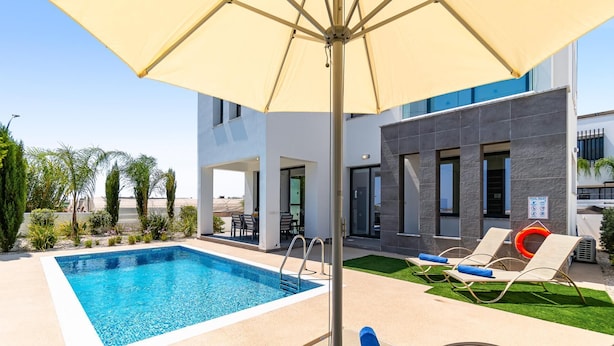 Ein Swimmingpool mit Liegestühlen und Sonnenschirm, umgeben von einem modernen Gebäude mit Poolbereich.