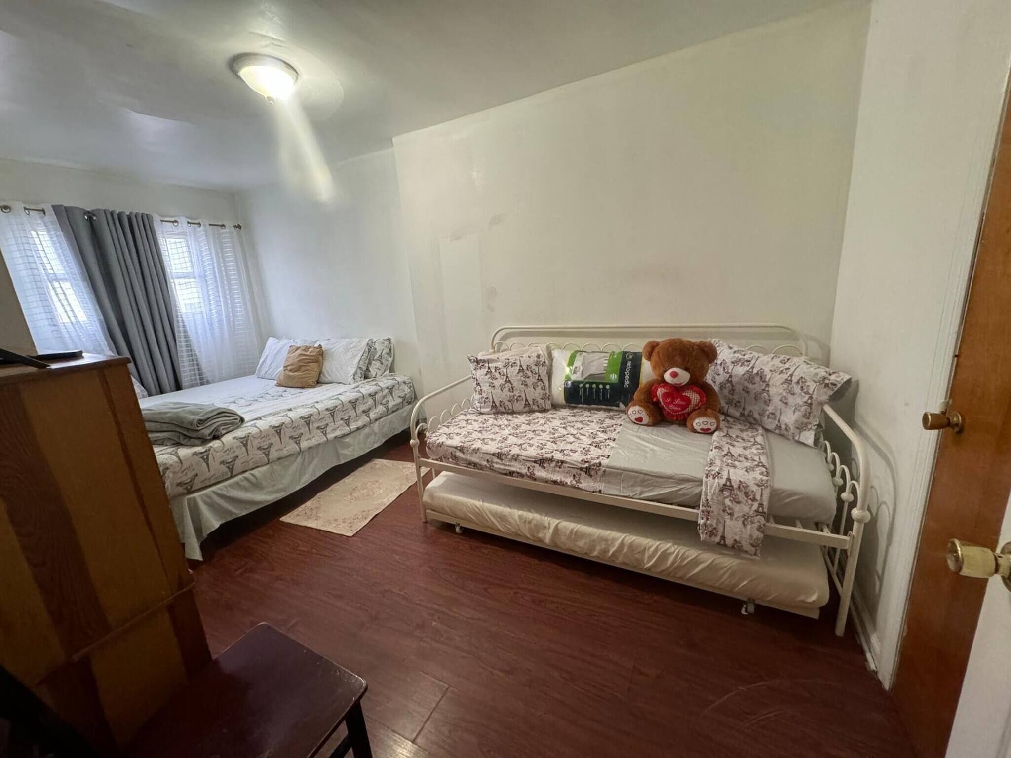 A small, single-room accommodation with two beds, a wooden floor, a teddy bear on one bed, and a door to the right.