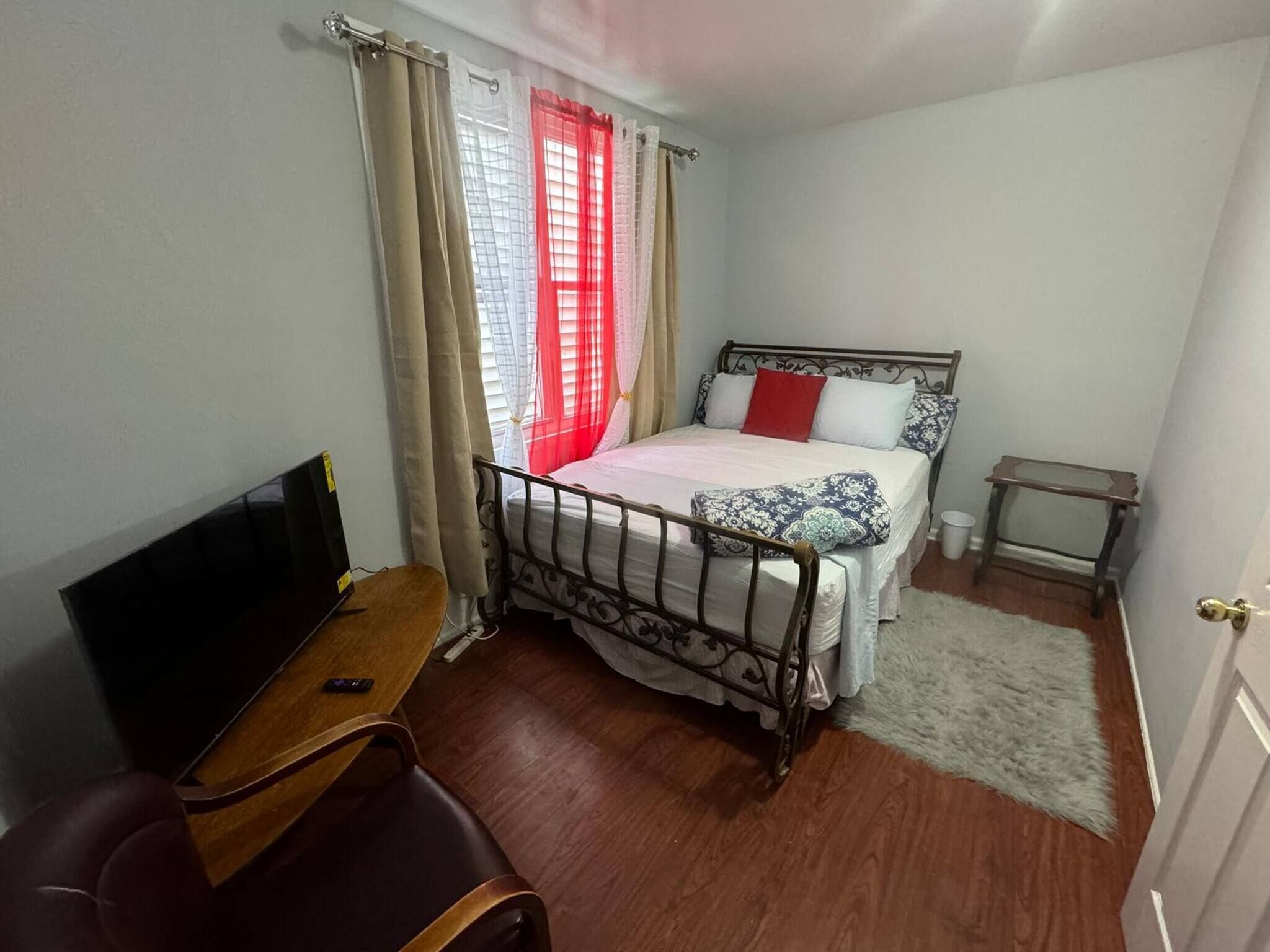 Une chambre à coucher avec un lit, une télévision, une chaise, une petite table et une fenêtre avec des rideaux.