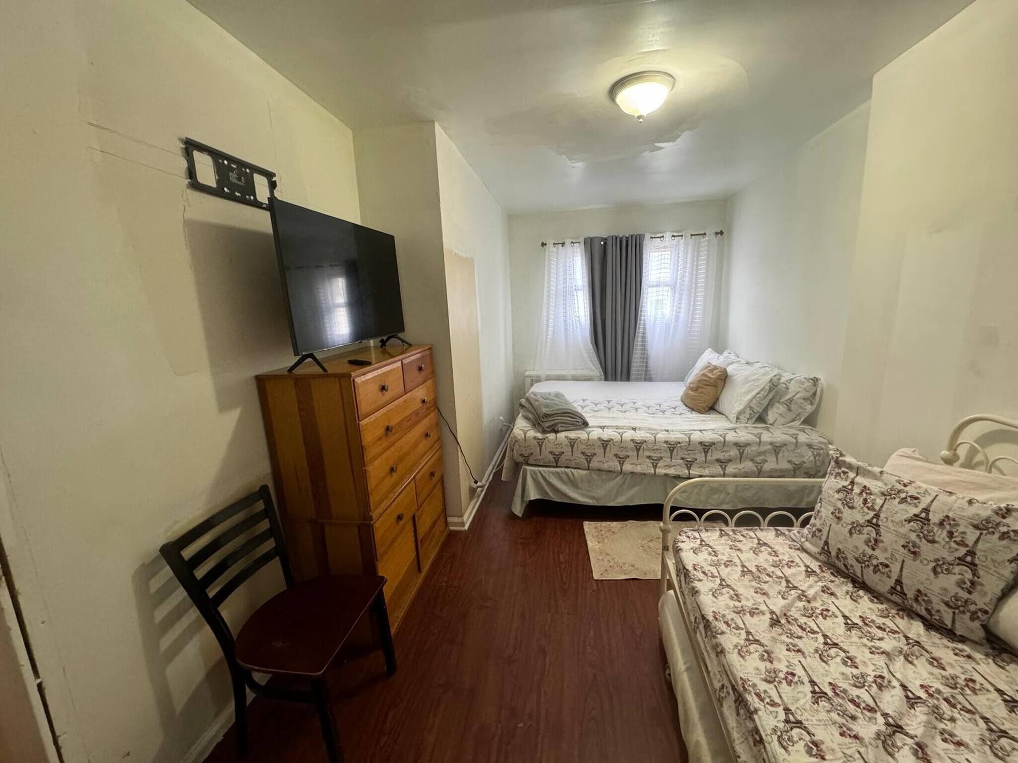 A bedroom with a wooden dresser, a television, a bed with floral bedding, a chair, and a window with curtains.