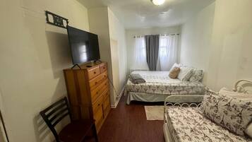 A bedroom with a wooden dresser, a television, a bed with floral bedding, a chair, and a window with curtains.