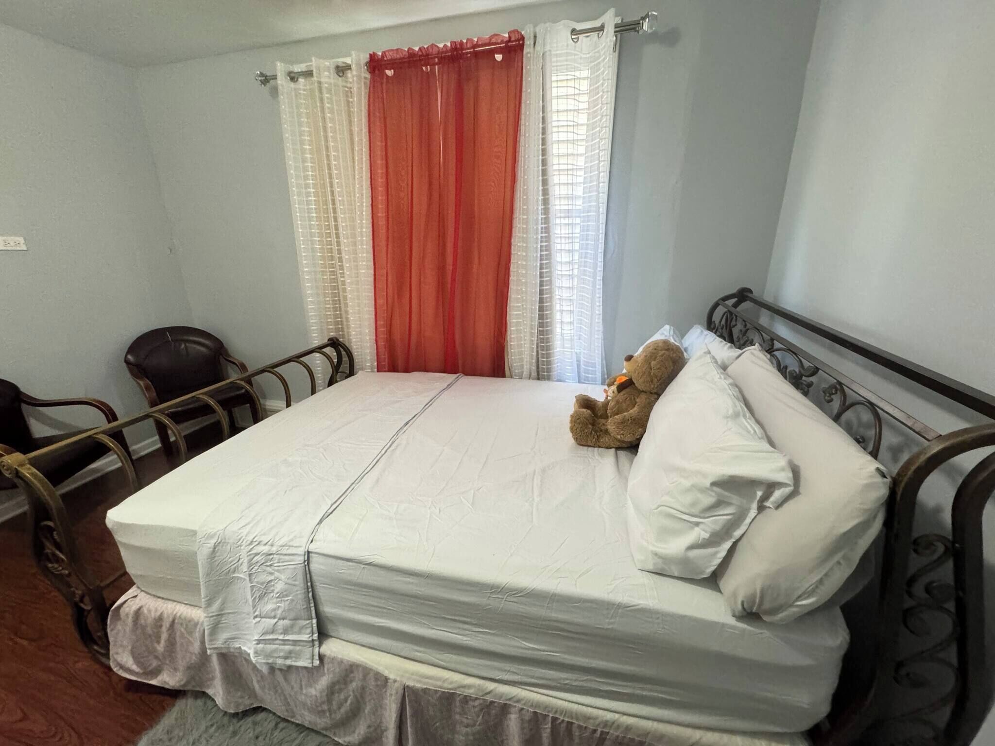 A single-bed room with a mattress, pillows, a teddy bear, a chair, and curtains.