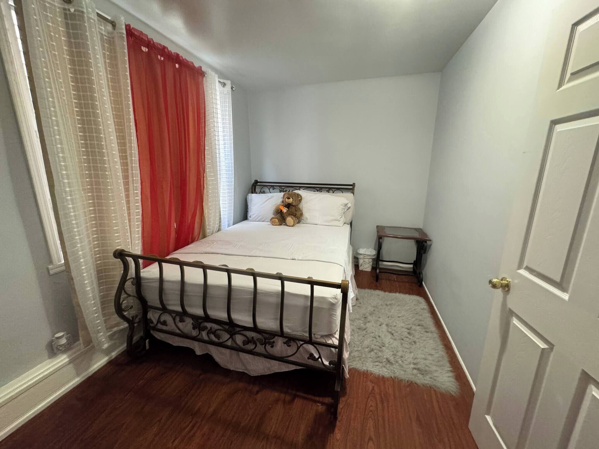 Un dormitorio con una cama, una mesita de noche pequeña, un osito de peluche y una ventana con cortinas.