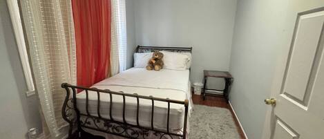 Un dormitorio con una cama, una mesita de noche pequeña, un osito de peluche y una ventana con cortinas.