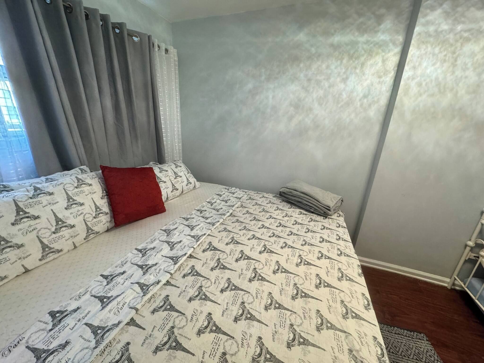 Quarto com uma cama, cortinas cinzas, um travesseiro vermelho e um edredom estampado.