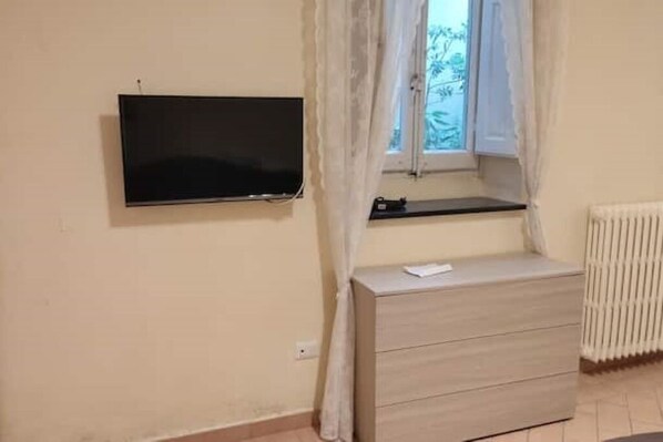 A room with a flat-screen TV mounted on the wall, a window with a curtain, and a wooden dresser with drawers. - Centralissimo: Comodo per 3, Economico per 6 :-) (Firenze)