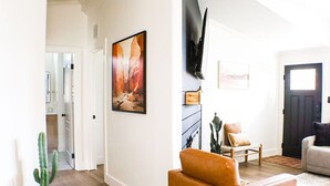 A living room with a sofa, a coffee table, and a TV mounted on the wall. There is a door leading to another room and a cactus plant in the corner. - Trail Chaser Moab, UT (Moab)
