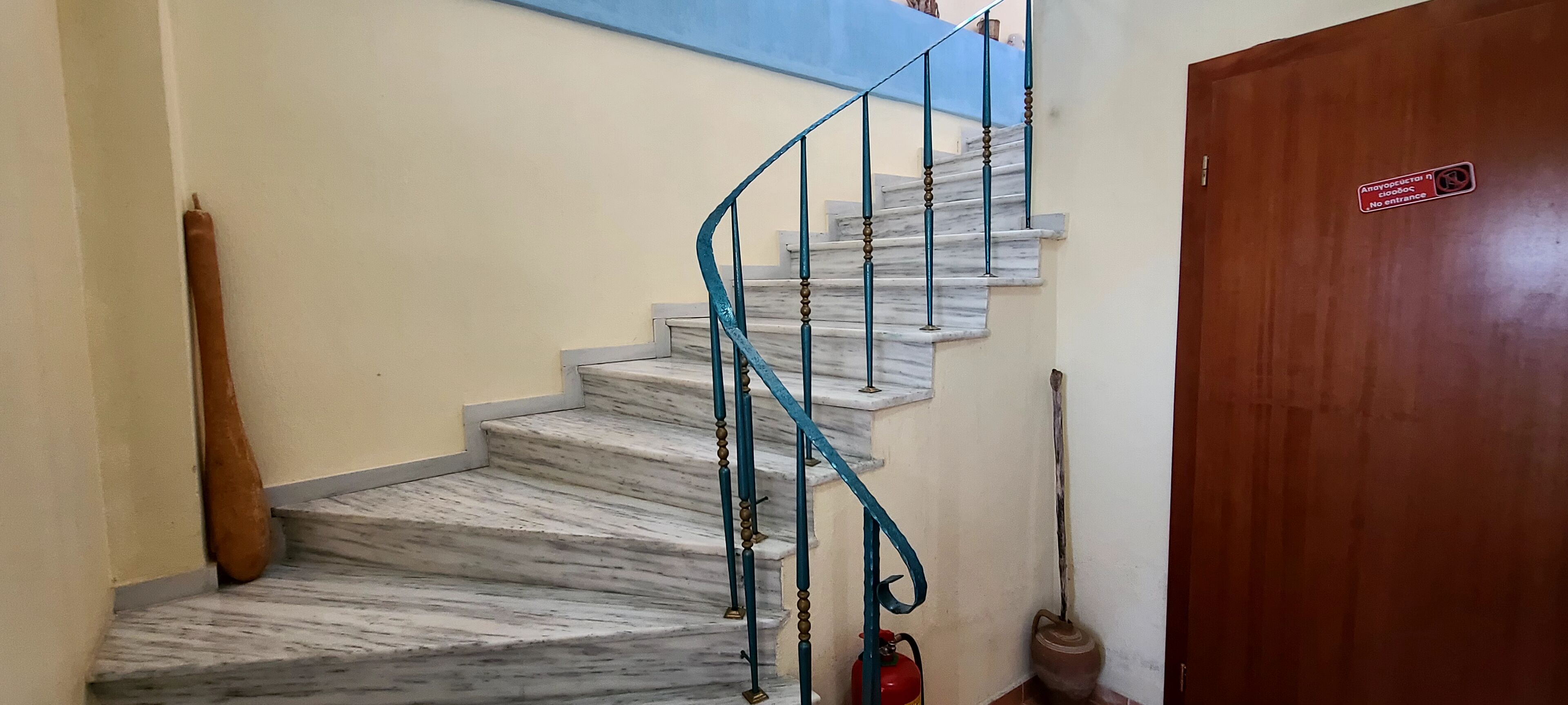 A marble staircase with a blue handrail, a red fire extinguisher, and a wooden door with a sign.