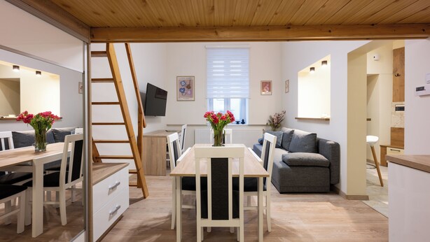 A modern interior with a wooden ceiling, a dining area with white chairs and a table, a grey sofa, and a kitchen area with a wooden countertop. - Native Apartments Nowowiejska 9 (Kraków)