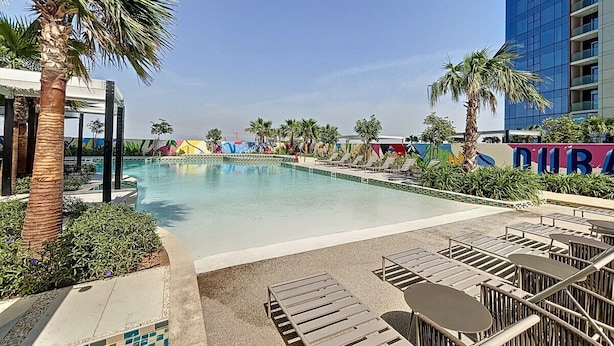 A swimming pool with lounge chairs and palm trees, surrounded by a modern building with a sign that reads "DUBAI". - Elegant 2 Bed| Business Bay | Central & Convenient - Aykon City Tower (Business Bay)