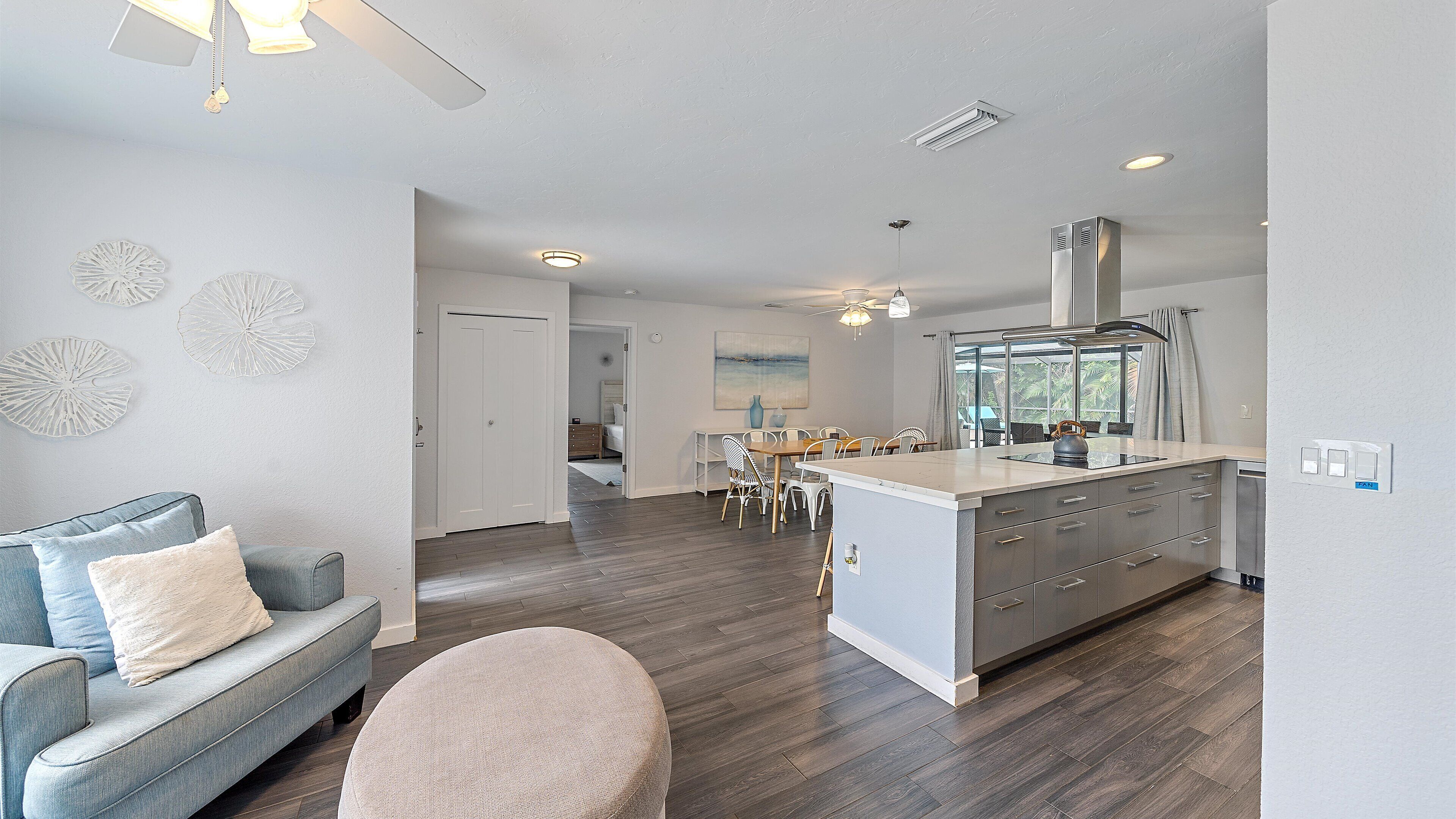 A modern kitchen with a central island, a dining area with chairs, and a living room with a sofa and ottoman.
