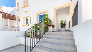 Una escalera conduce a la entrada de un edificio con fachada blanca, detalles amarillos y plantas en macetas.