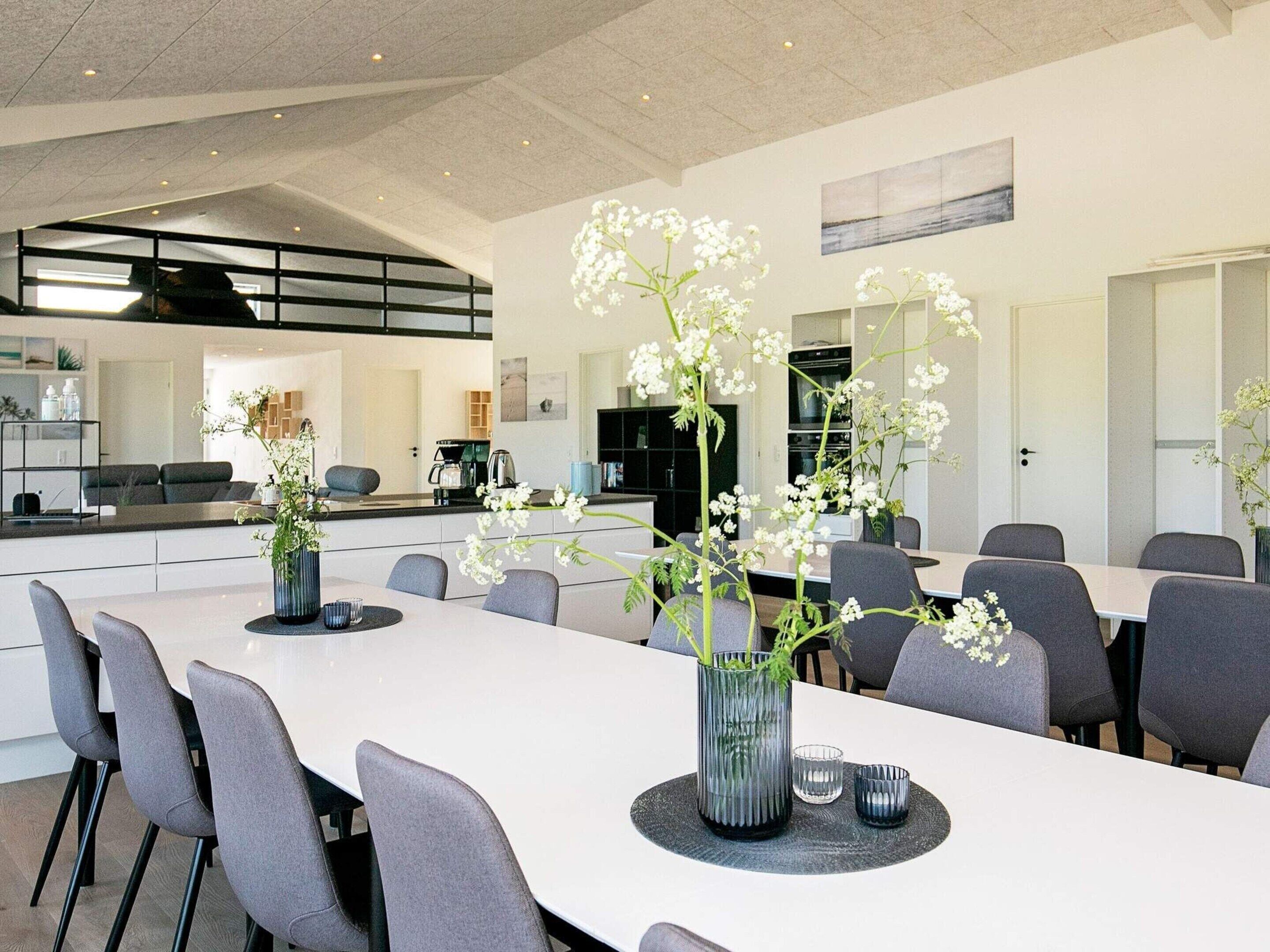 Un espace de salle à manger moderne avec une longue table, des chaises grises et des fleurs blanches décoratives disposées dans des vases.
