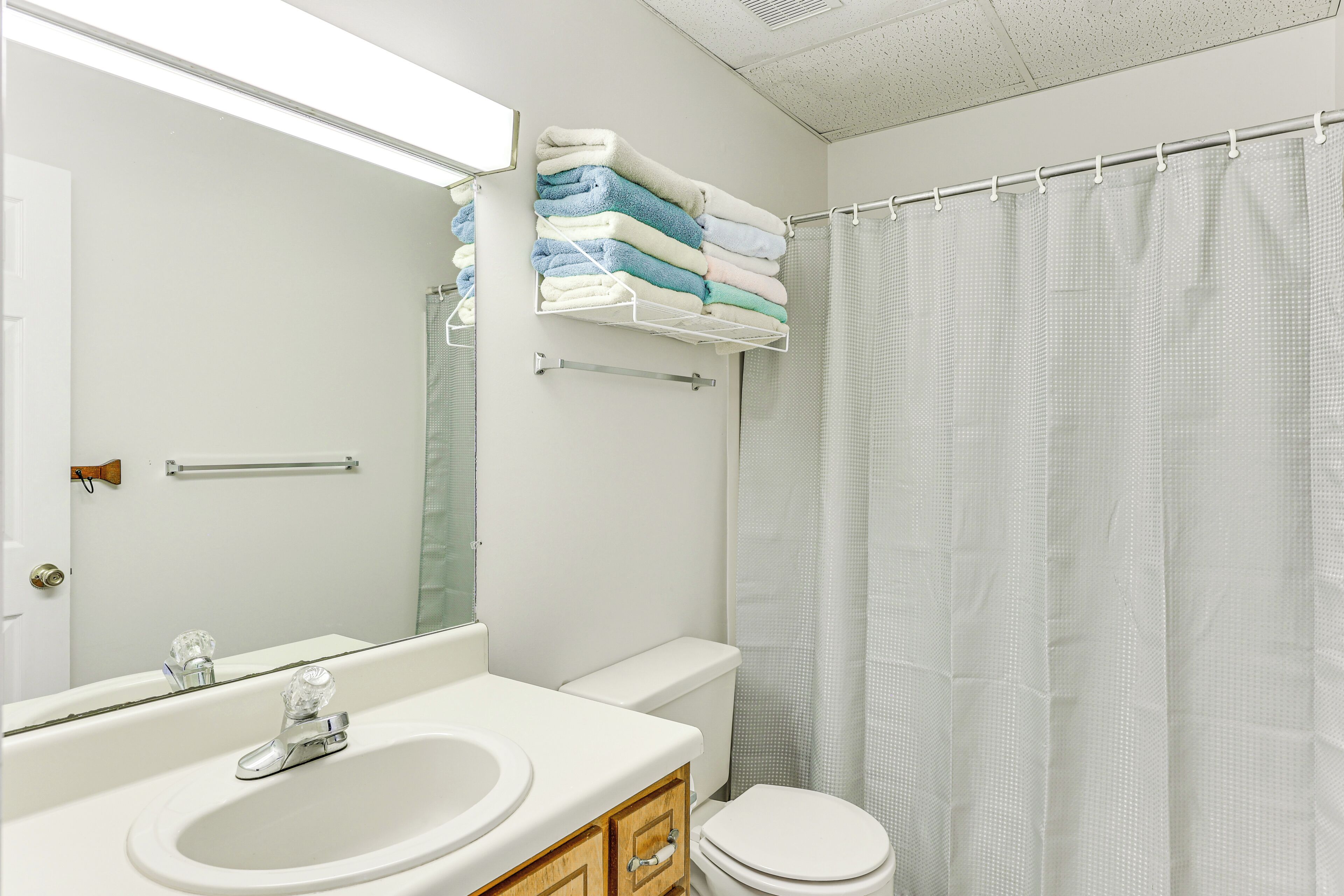 Une salle de bain avec un lavabo blanc, un miroir et des toilettes. On y trouve un porte-serviettes avec des serviettes pliées et un rideau de douche.